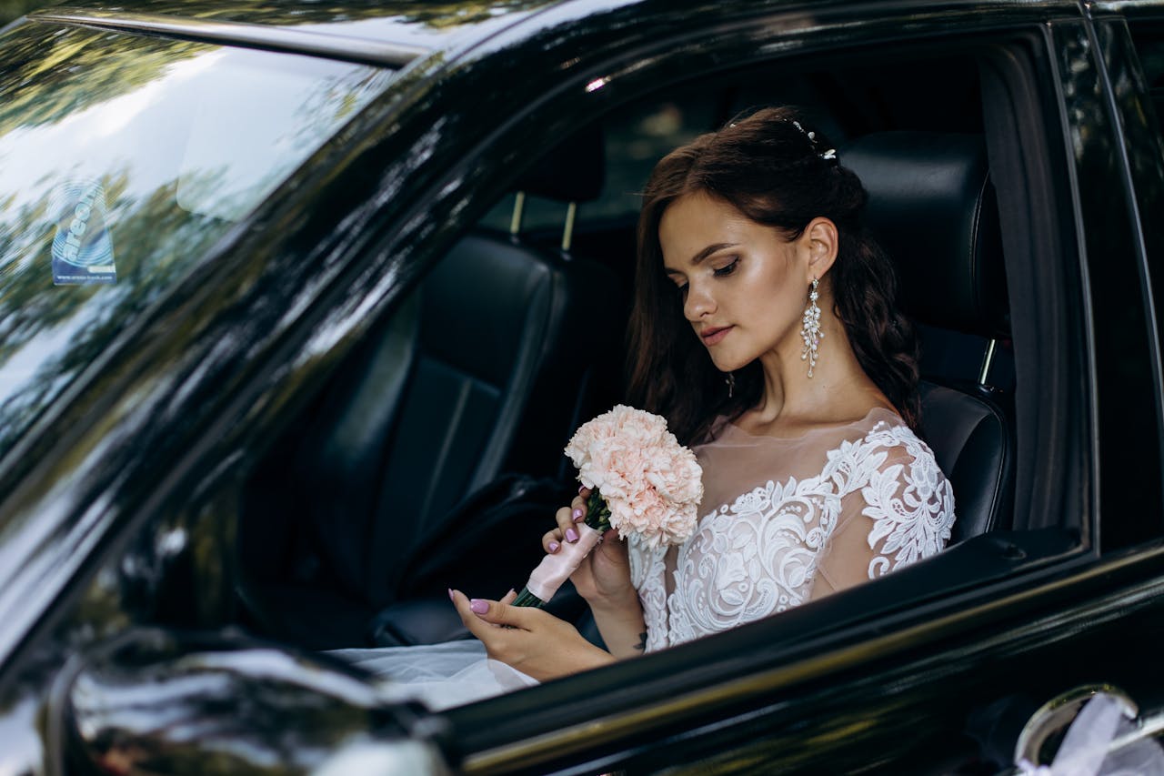 A bride wearing a lace wedding dress holds a bouquet sitting in a car, ready for her wedding celebration.